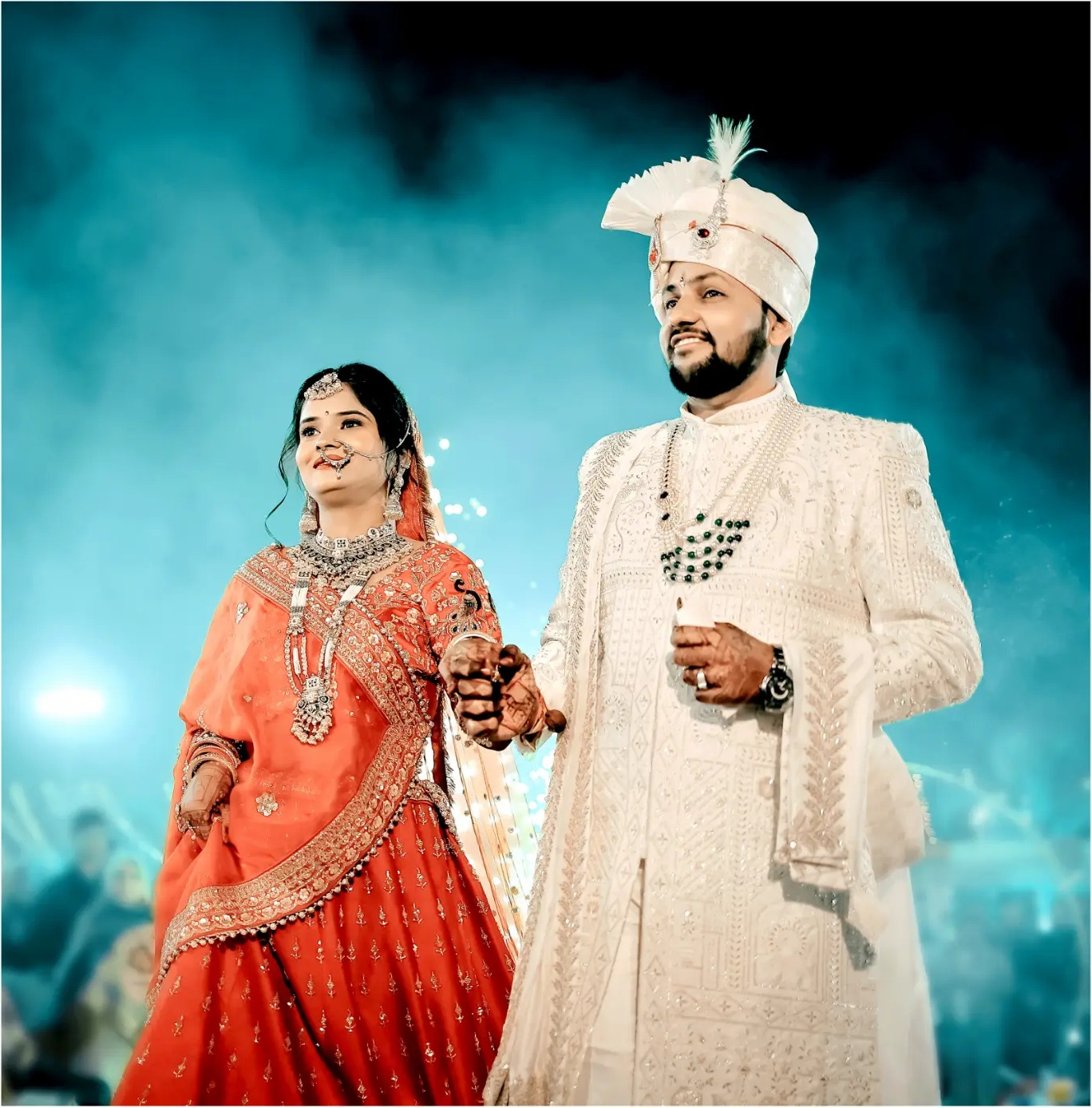 Bride in red lehenga and groom in white sherwani against teal stage lights